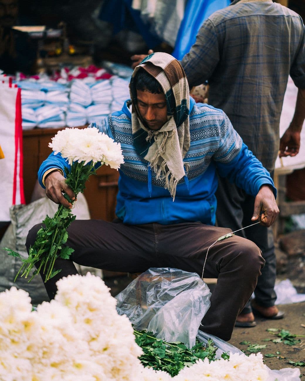 A flower seller in a knitted jumper and scarf tying white chrysanthemums into a bunch at the market