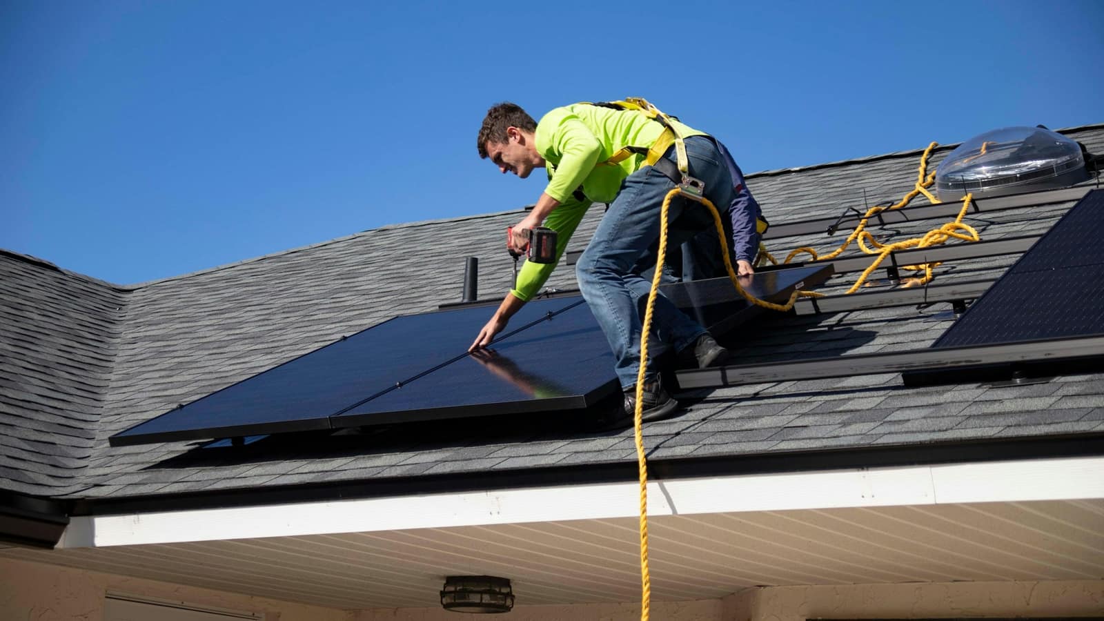 An installer on a safety line drilling a panel rail into place on a sloping roof