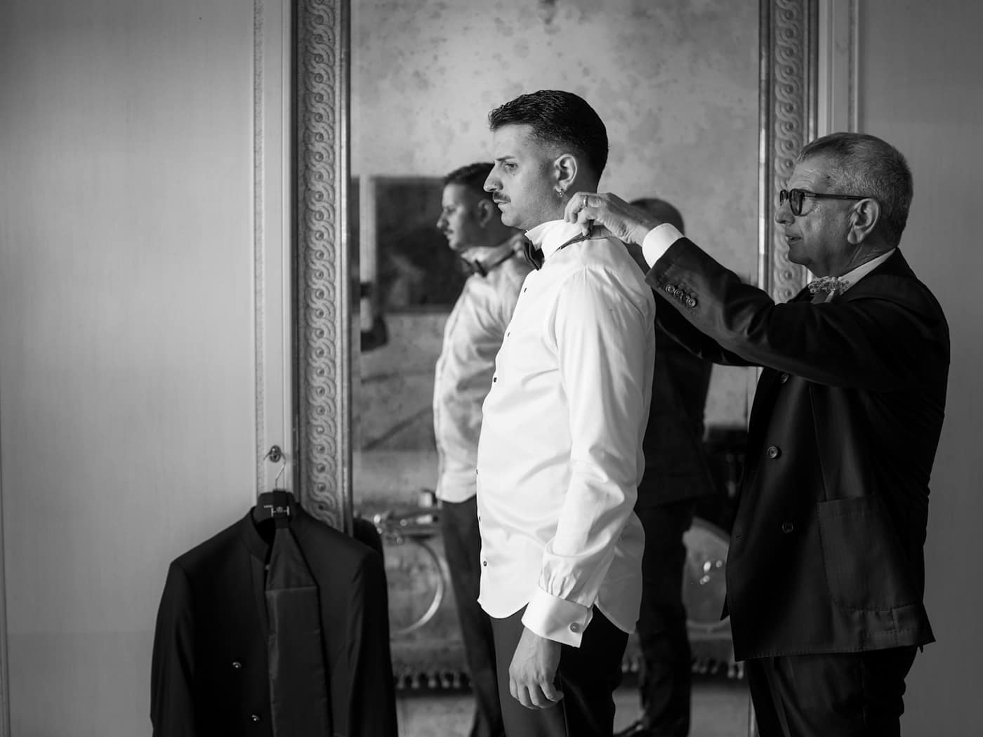 An older tailor setting the shirt collar of a young client before his jacket goes on