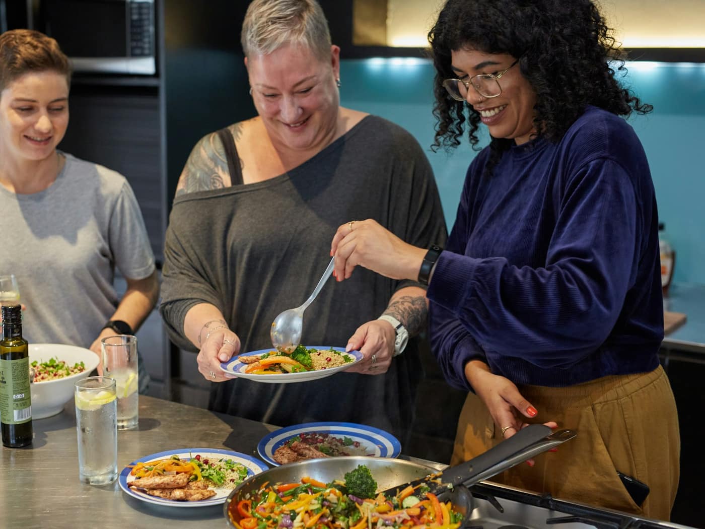 A cook and two students smiling as they plate a pasta dish in a bright kitchen