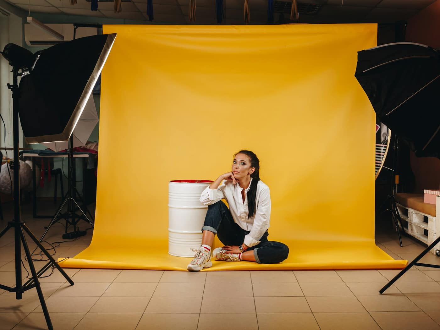 A photographer working beside a softbox while a client sits on a stool against a yellow backdrop