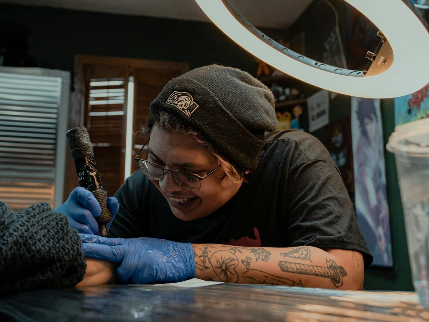 A tattoo artist in a beanie smiling as he works on a client's forearm