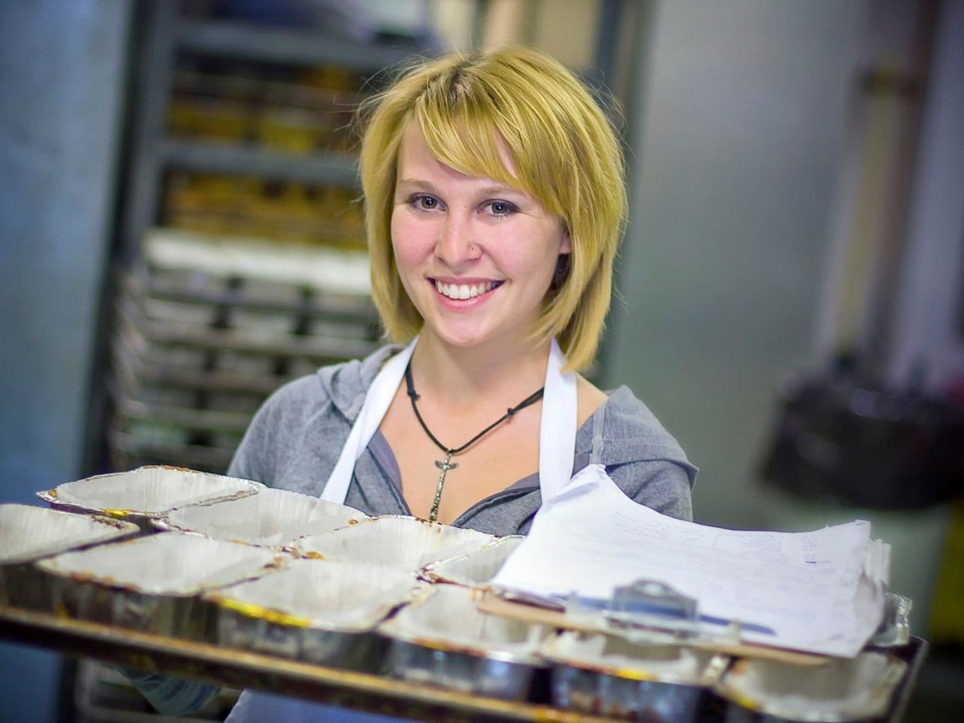 A smiling baker in an apron holding a tray of pastries in a bakery kitchen