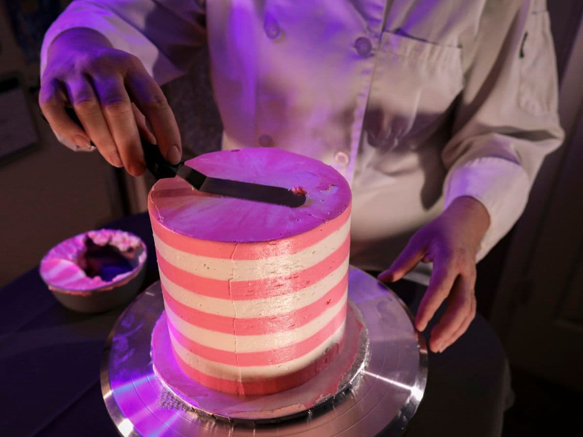 A pink and white striped cake on a turntable, being smoothed with a knife