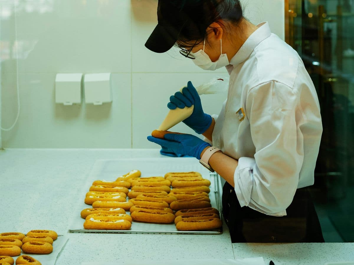A pastry cook in a cap piping onto a tray in a tiled kitchen