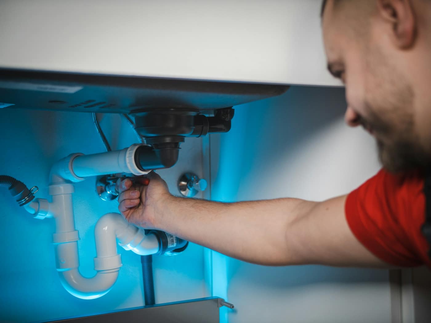 A plumber in a red shirt fitting the waste pipe under a kitchen sink