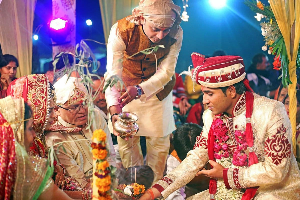 A groom in a red turban performing a ritual with elders at a lit stage