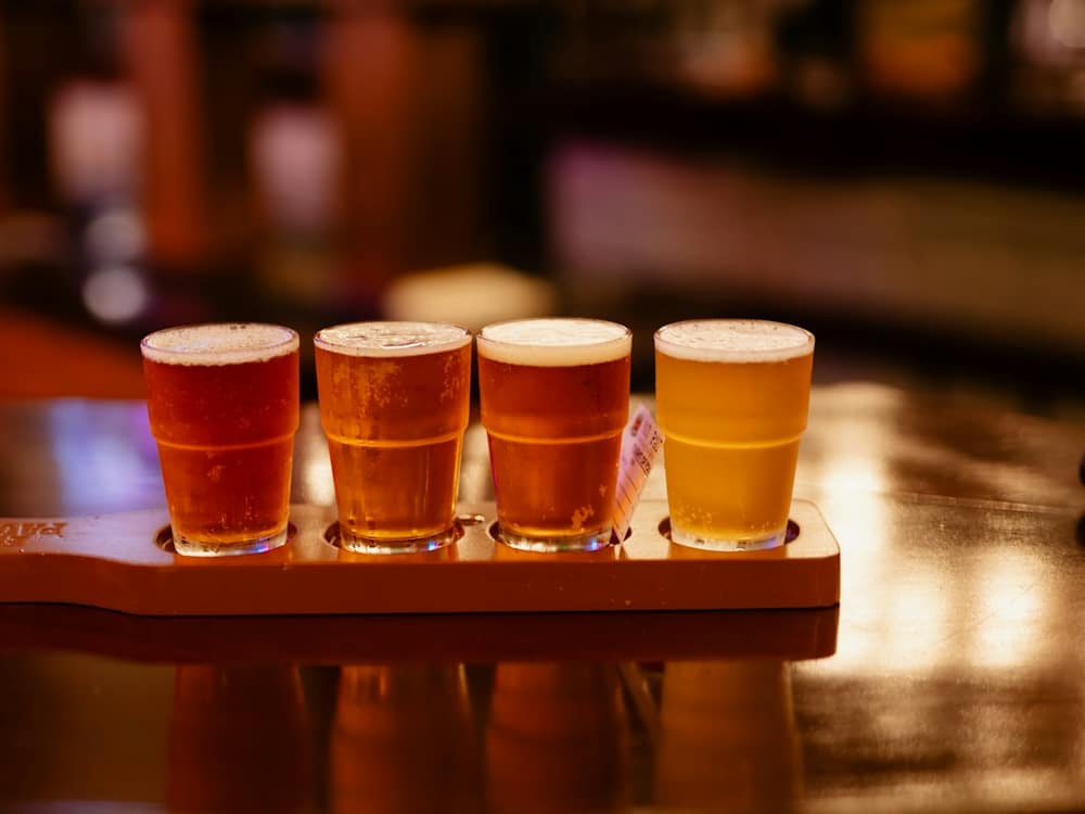A flight of four small glasses of red-amber beer on a wooden paddle