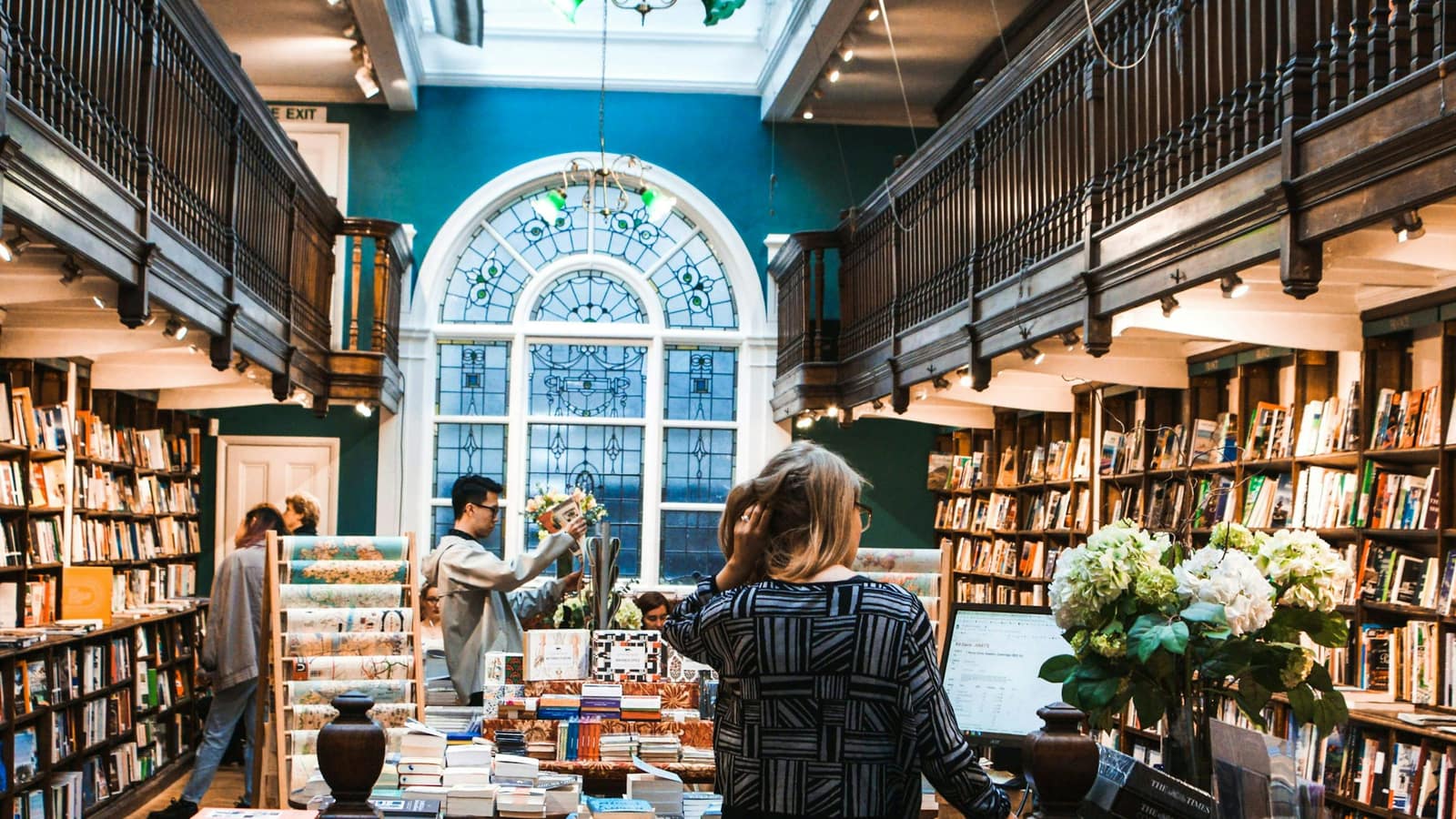A bookshop with an arched window, shelves to the ceiling and a bookseller at the counter