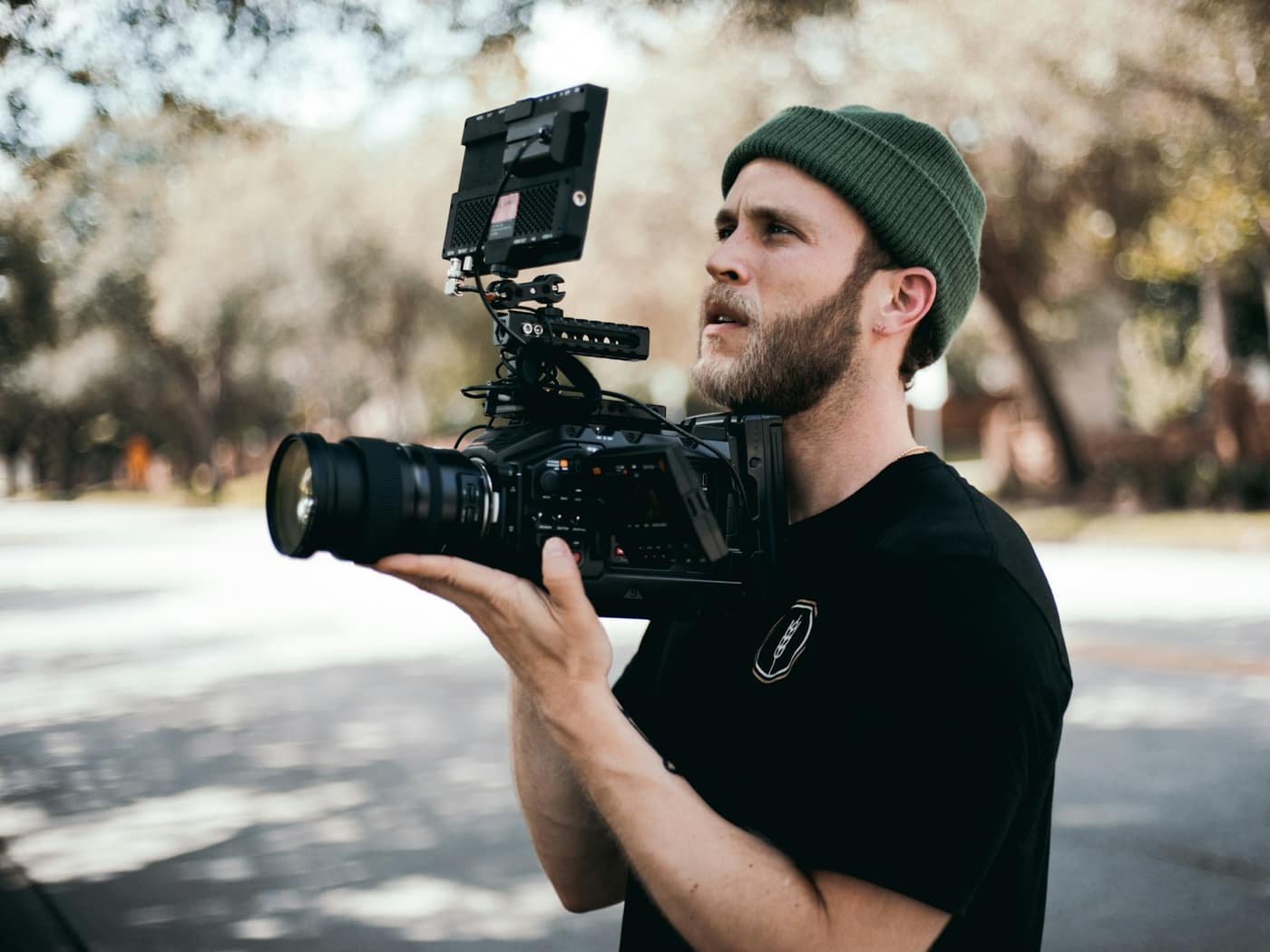 A cameraman in a beanie holding a cinema camera rig outdoors under trees